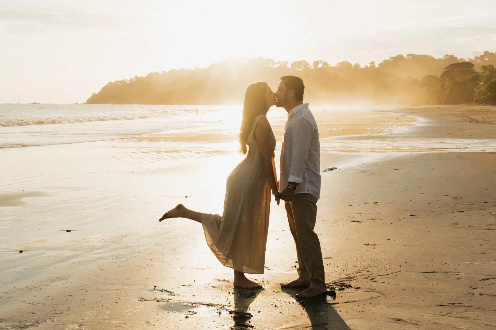Una pareja se besa a la orilla del agua en la playa frente a Miamar a la hora dorada, con el sol brillando detrás del promontorio boscoso.