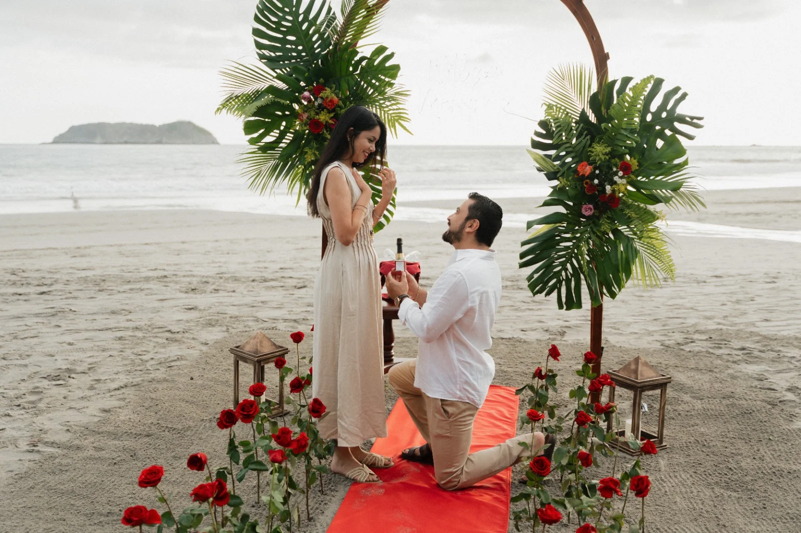 Un hombre de rodillas pide matrimonio bajo un arco floral de hojas de palma y rosas rojas en la playa de Miamar, con el Pacífico y un islote rocoso detrás.