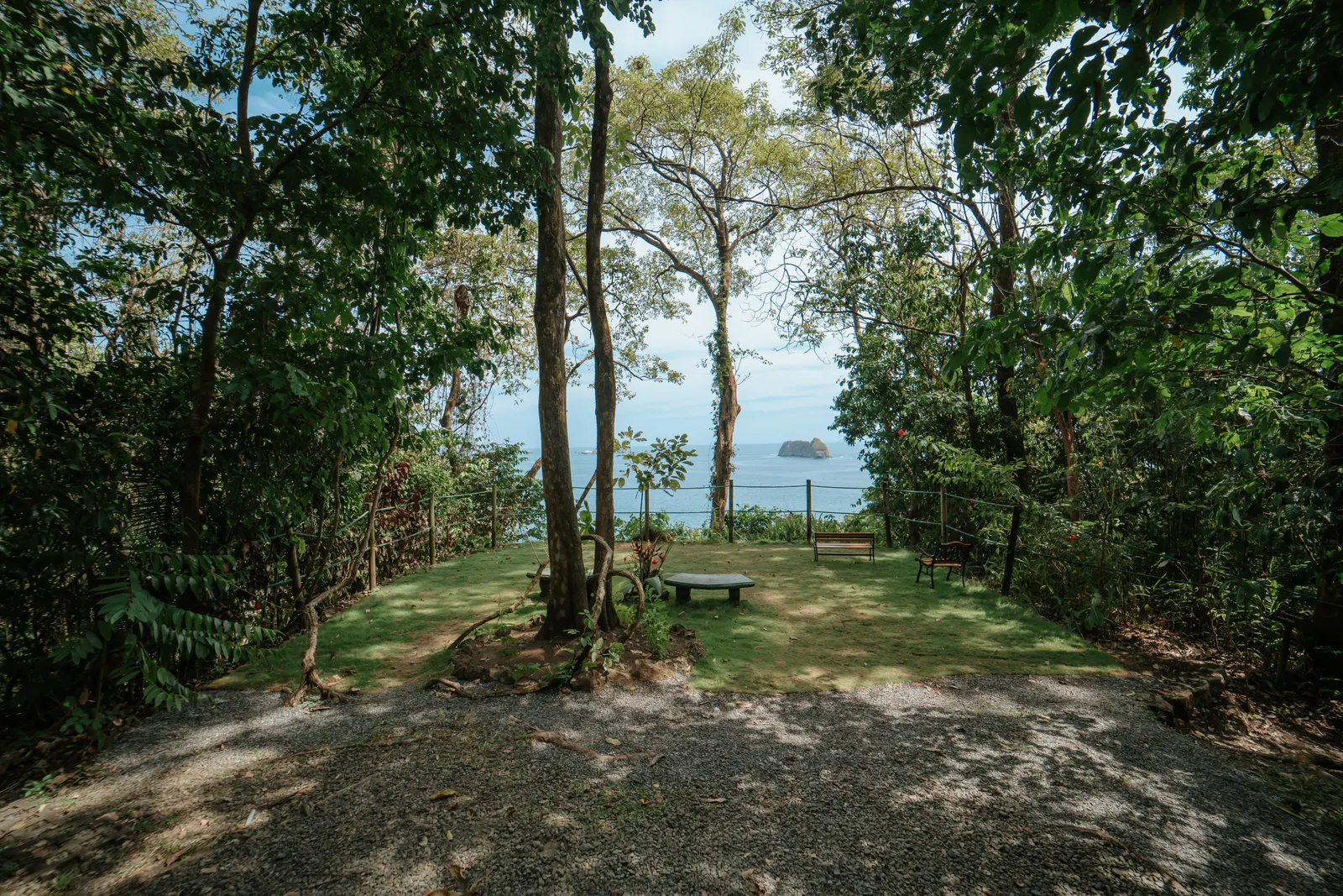 Vista amplia de un claro con mirador en el jardín enmarcado por árboles altos, con un sendero de lastre que entra y bancas al fondo con el océano más allá.