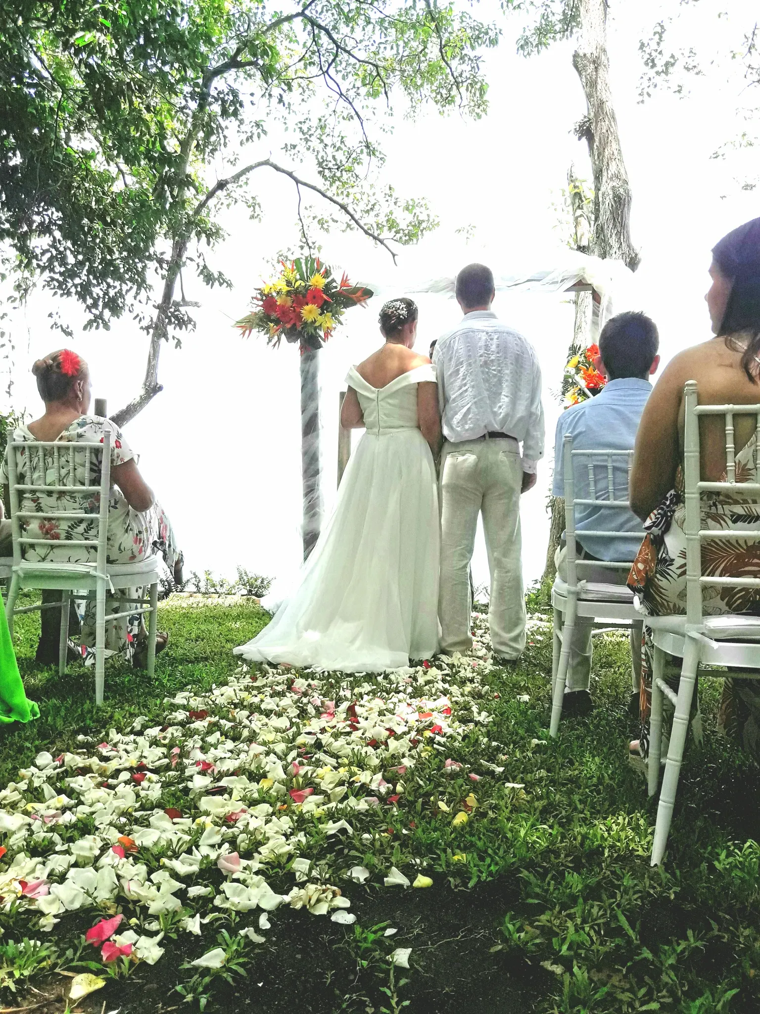Una pareja de espaldas frente a un arco floral sobre un césped cubierto de pétalos, con invitados sentados cerca y el océano al fondo.