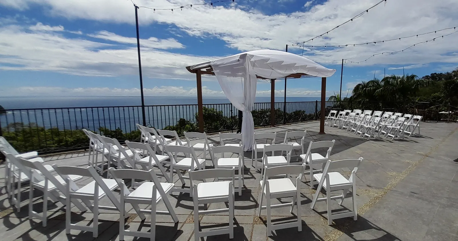 Montaje de ceremonia de boda al aire libre en Parador Nature Resort con sillas plegables blancas y un arco con telas translúcidas frente al océano Pacífico.