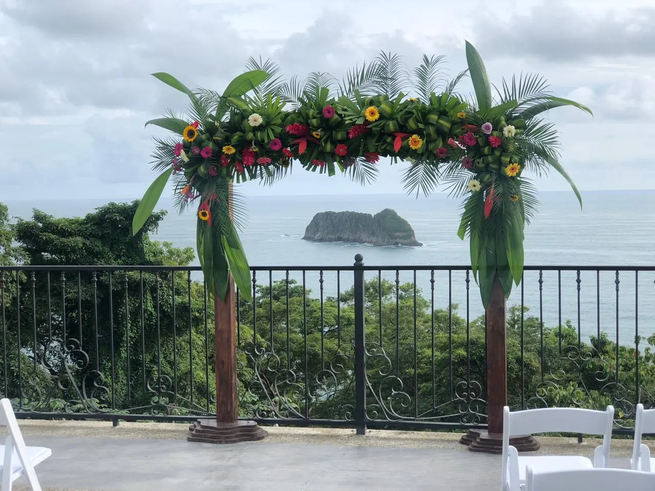 Arco de boda con flores tropicales y hojas de palma en una terraza, con un islote rocoso en el mar y sillas plegables blancas al frente.