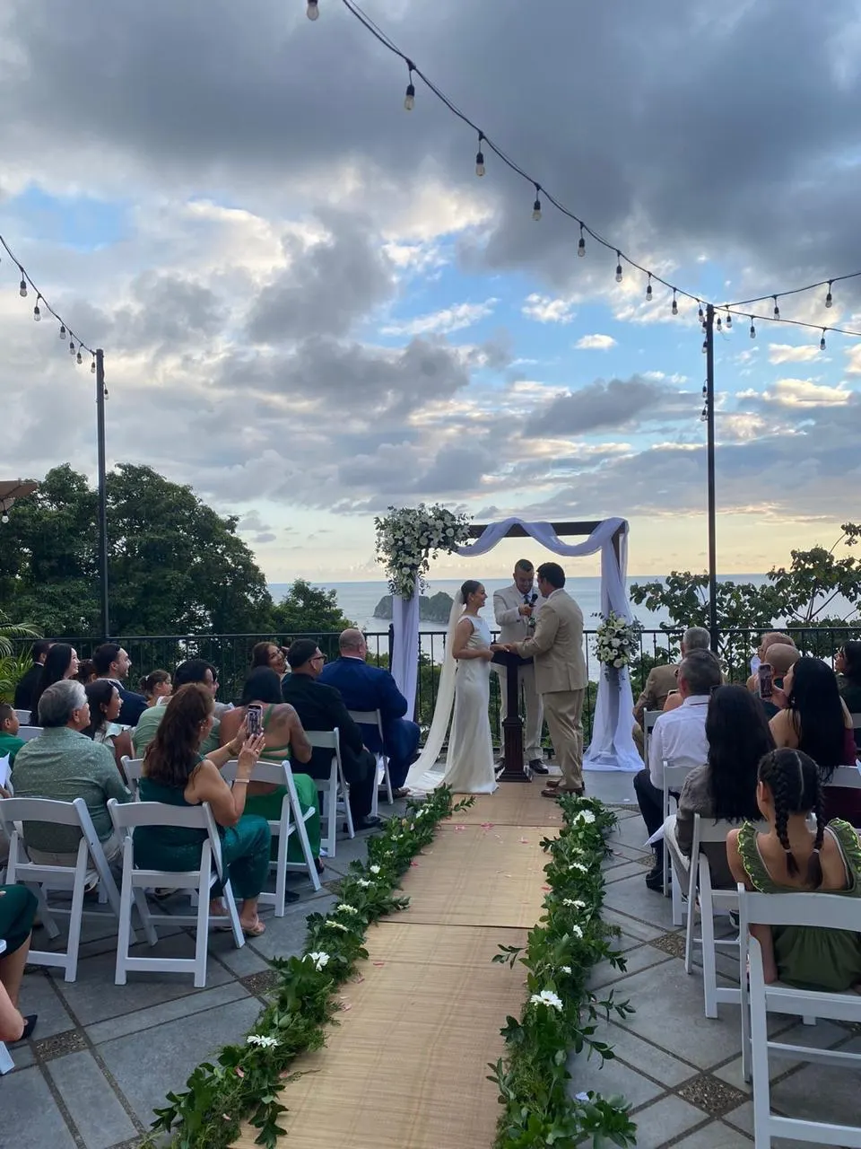 Los novios intercambian votos bajo un arco con telas blancas en una terraza sobre el acantilado con vista al océano al atardecer, con los invitados sentados a lo largo de un pasillo con flores.