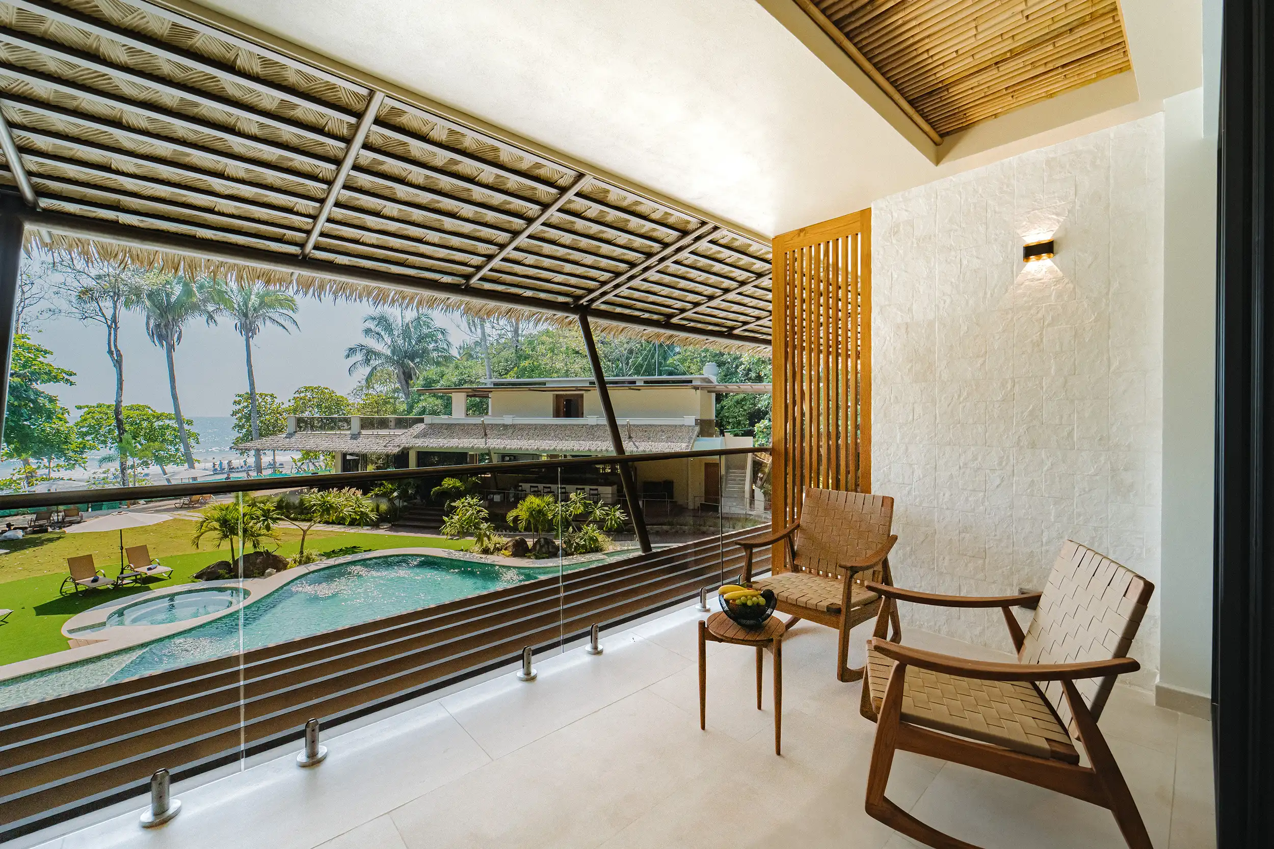 miamar-suite-2nd-floor-pool-balcony-hl23 Balcony seating area with woven wooden chairs along a glass railing, overlooking a pool and tropical palm trees.