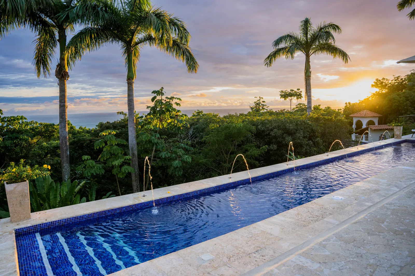 An infinity pool with fountain jets glows orange and pink at sunset, framed by palm trees and hills.