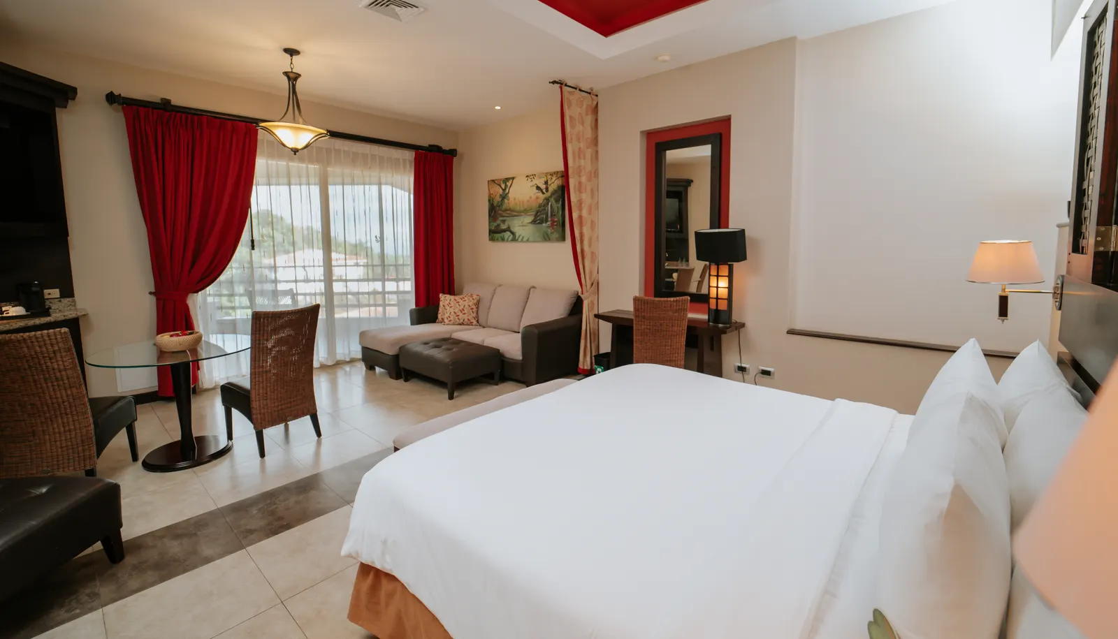 King bed and sitting area with sofa and glass table in the Parador Master Suite, balcony with red curtains open to a hillside view.