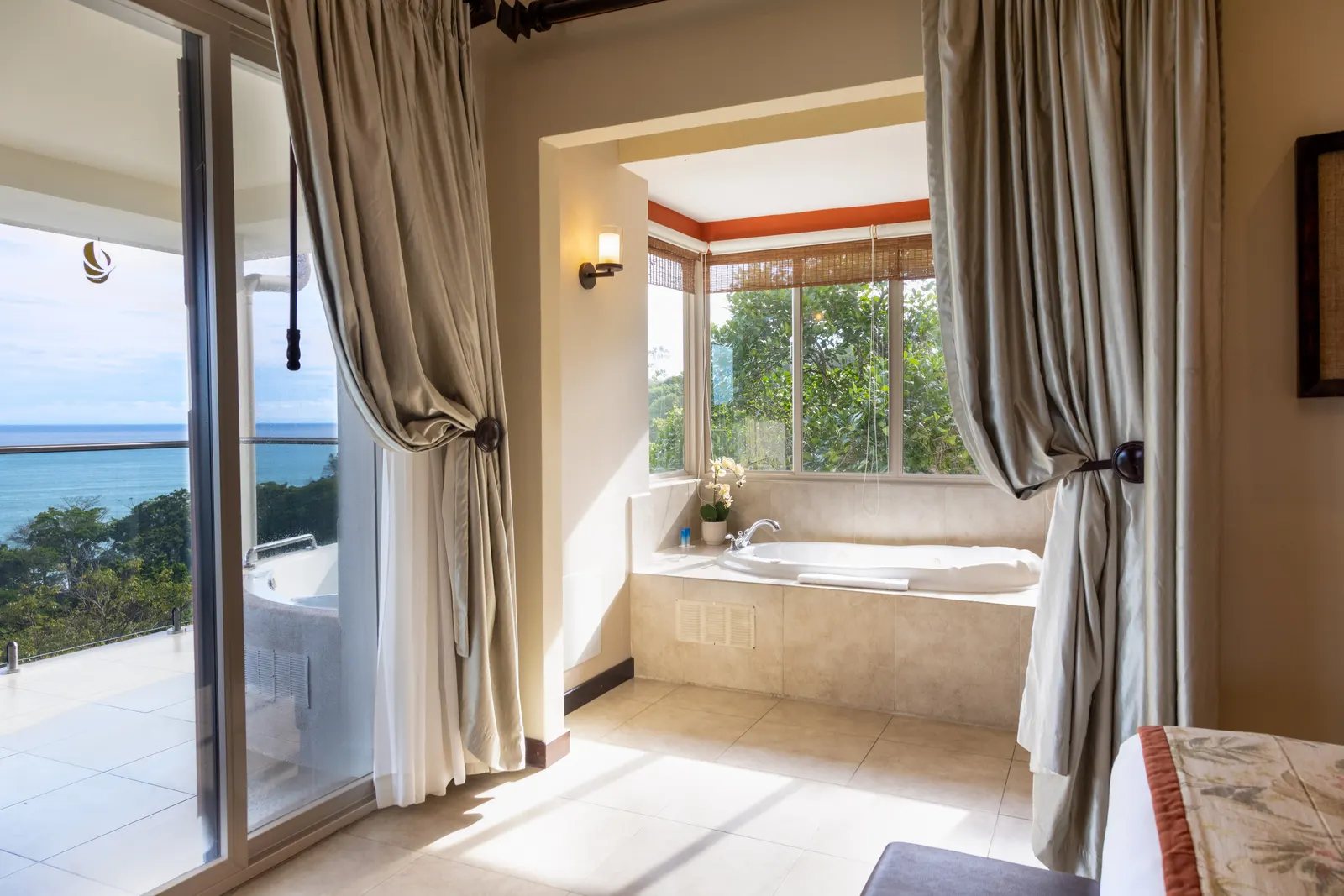 A jacuzzi tub in a tiled bay window nook with an orchid on the ledge, beside a sliding glass door open to an ocean-view terrace.