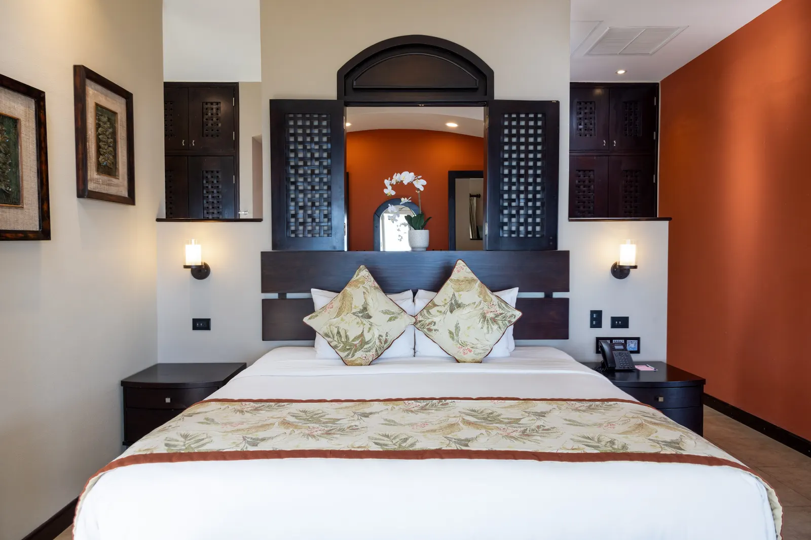 A king bed with floral linens beneath a dark wood alcove with open mirrored shutters and an orchid, beside an orange accent wall.