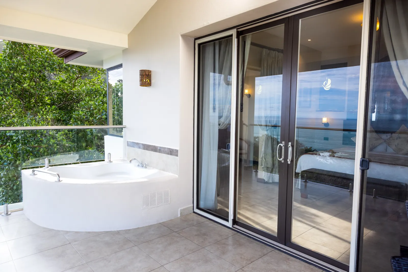 A private terrace with a round jacuzzi tub, a glass railing, treetops, and sliding glass doors reflecting the ocean.