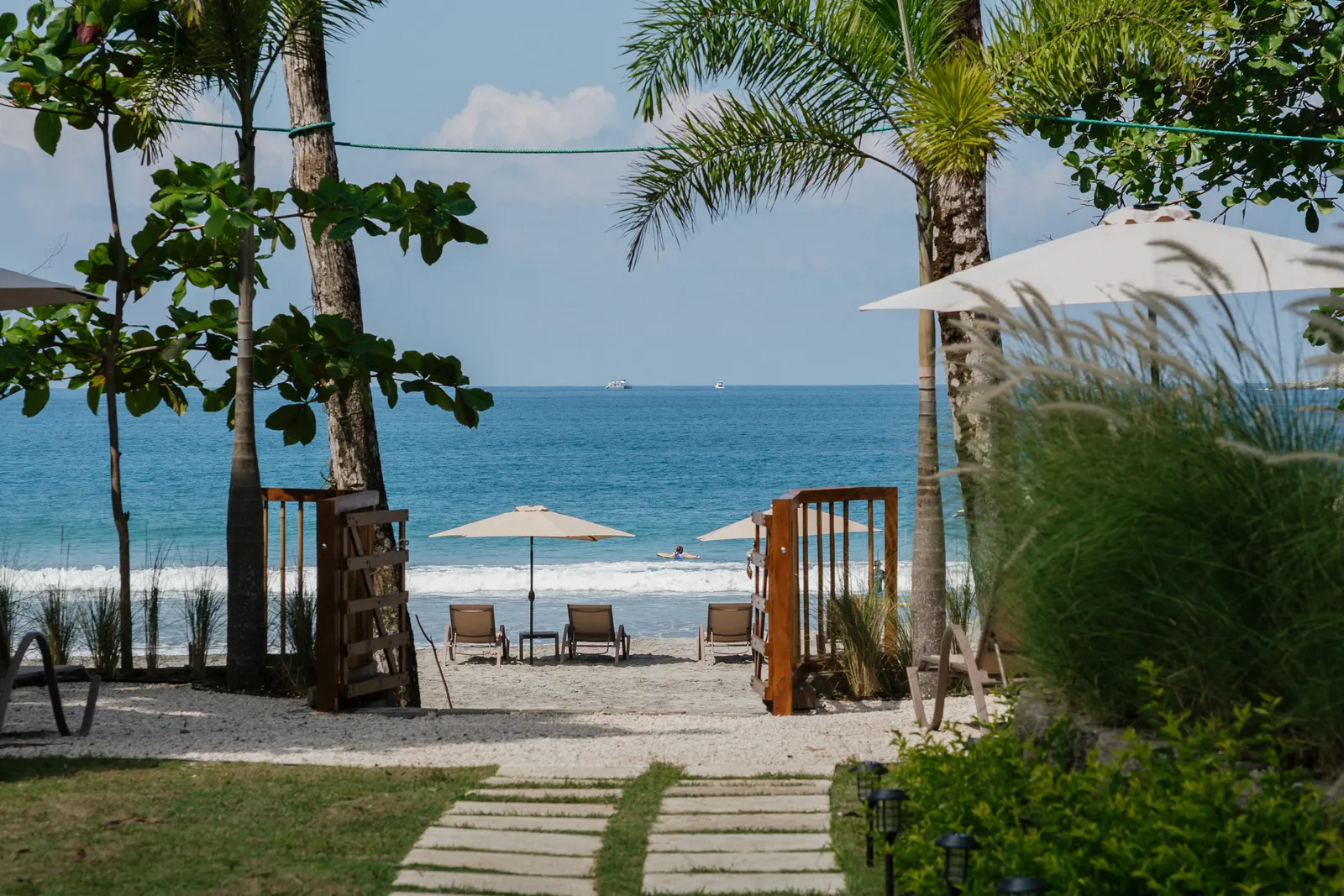 Wide view of a wooden beach gate and palm trees framing loungers on the sand, with a swimmer in the ocean waves beyond.