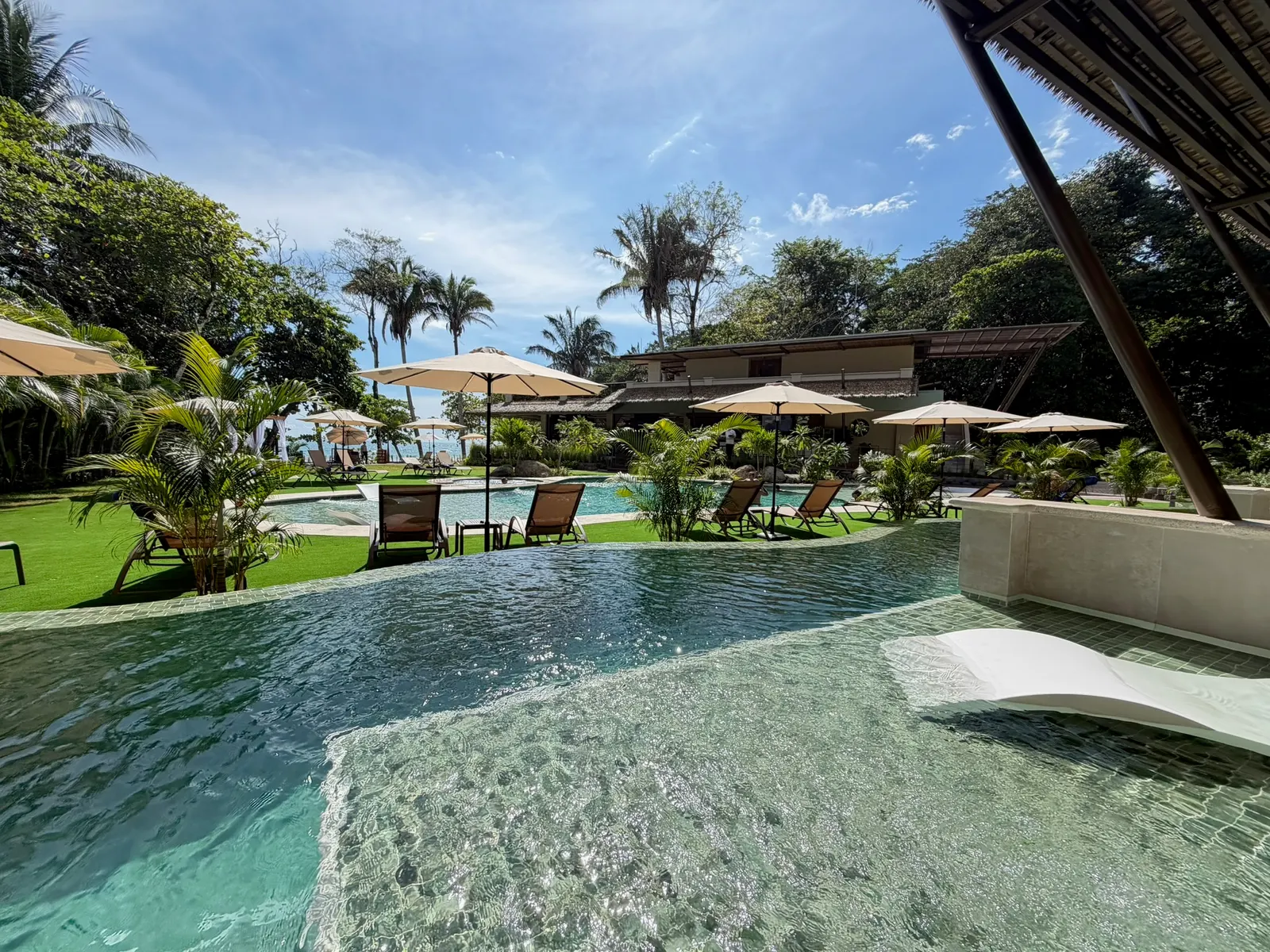 Infinity pool edge with lounge chairs, cream umbrellas, and the main building framed by palm trees under a clear blue sky.