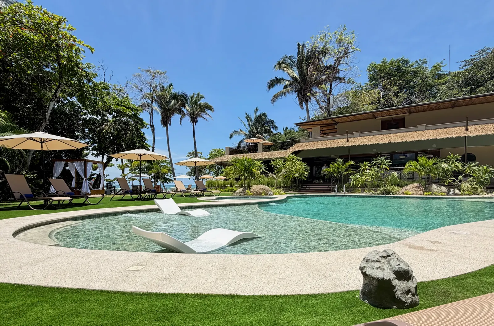 White manta-shaped loungers floating in the resort pool, with lounge chairs, umbrellas, and the thatched-roof building in the background.