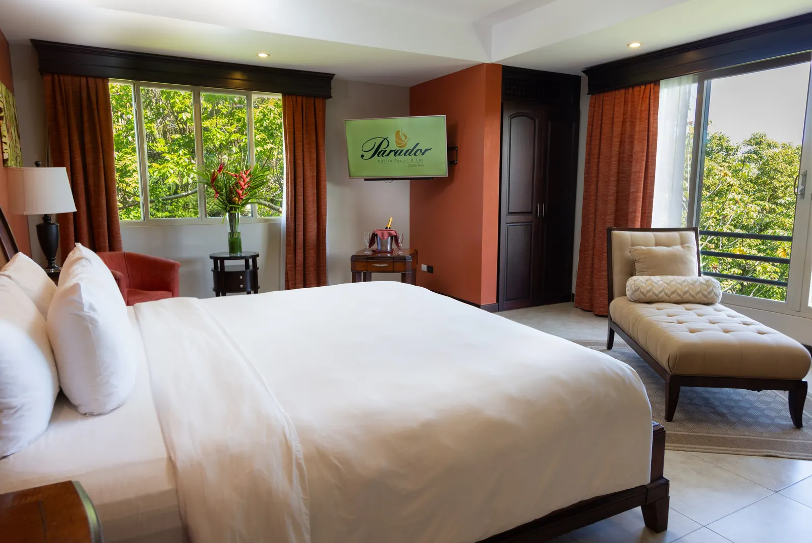 Bedroom with white bedding, a television showing the Parador logo, a red armchair, and a cream chaise lounge near a window.