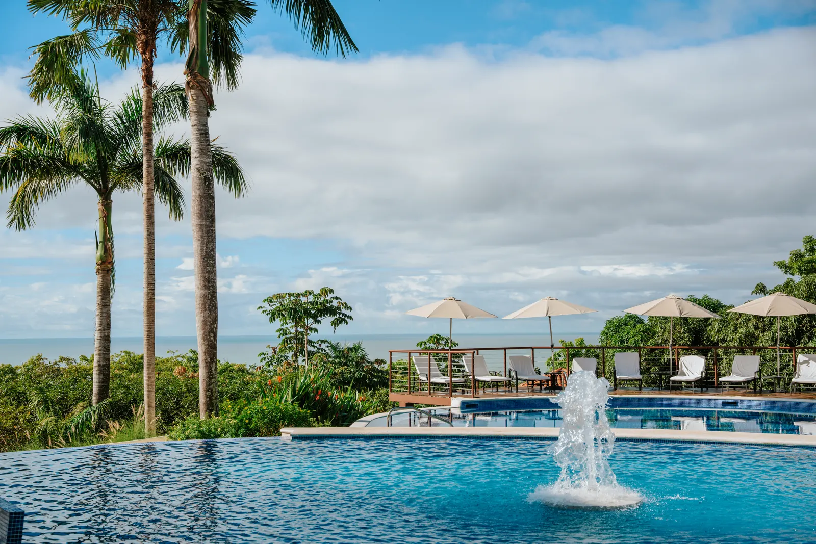 Lounge chairs and umbrellas beside the pool deck with a fountain jet and the ocean beyond.