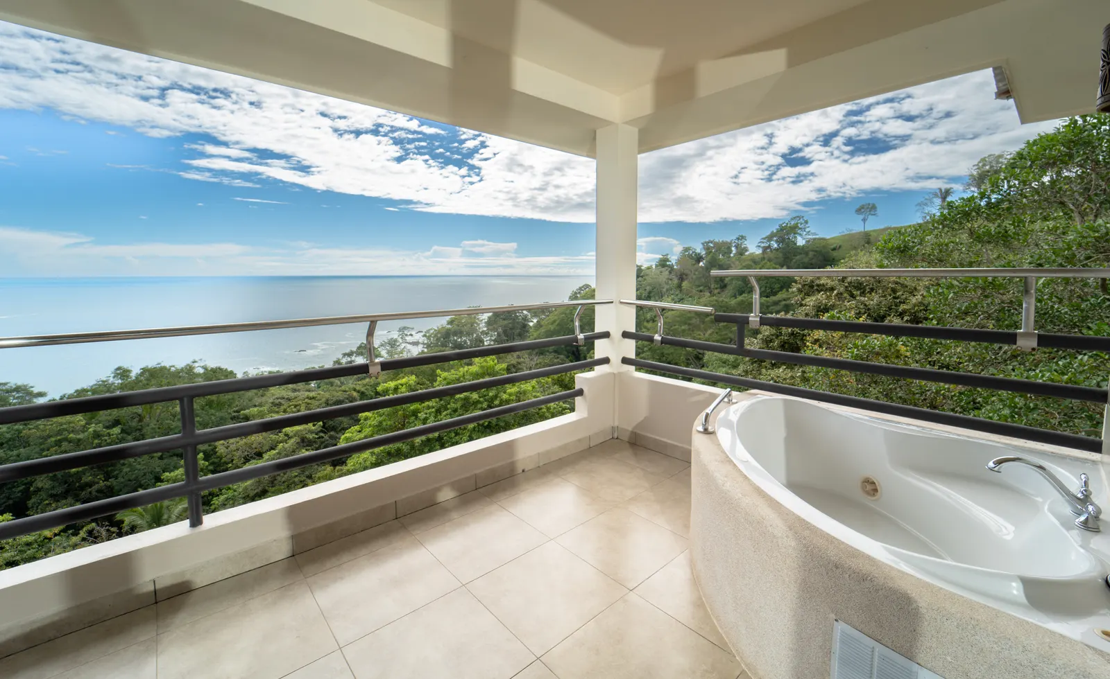 A wide hotel suite balcony with a railing overlooks a forested coastline and the ocean under a blue sky.