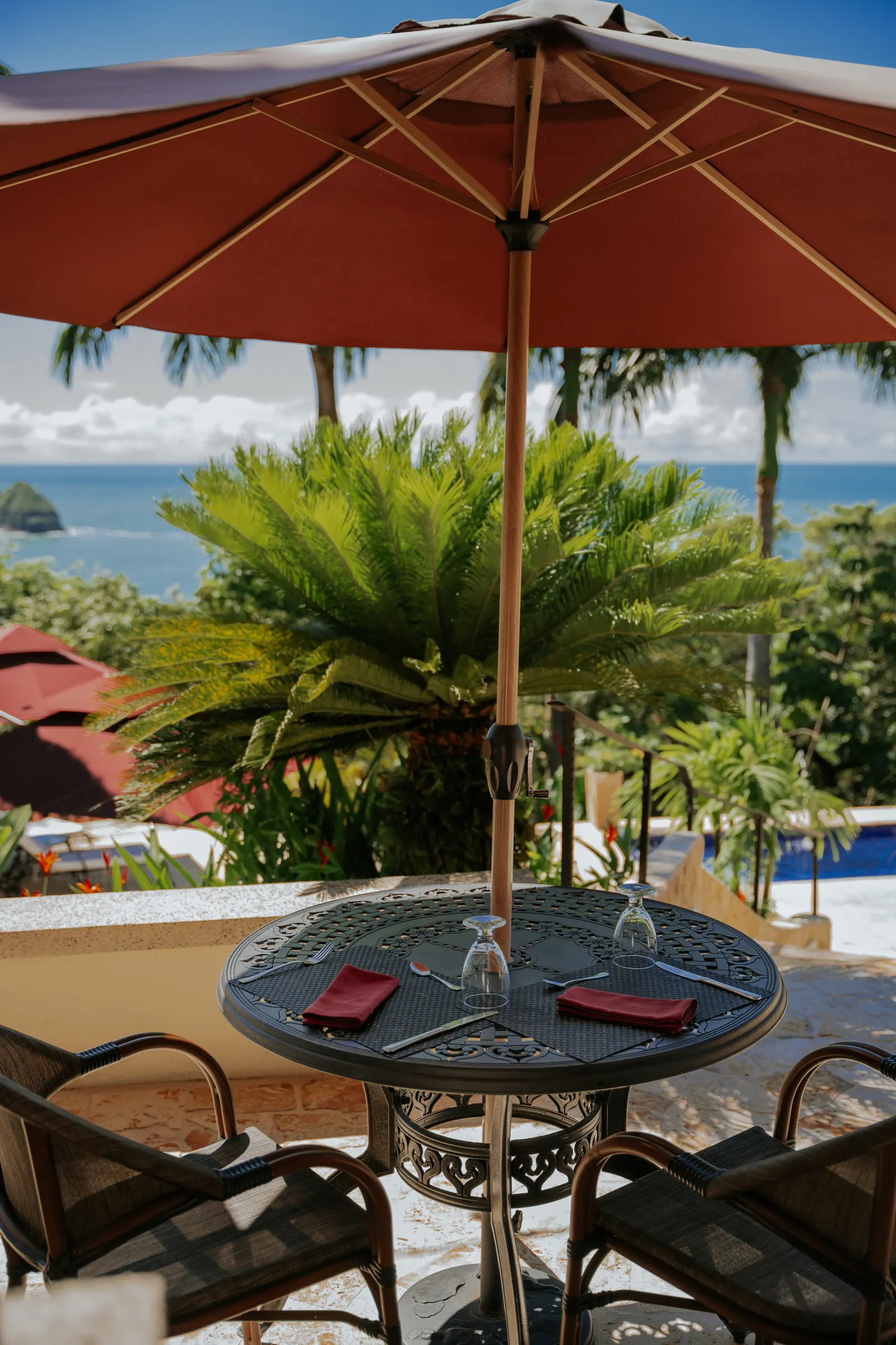 A terrace dining table set for two under a red umbrella, with the pool and ocean in the background.