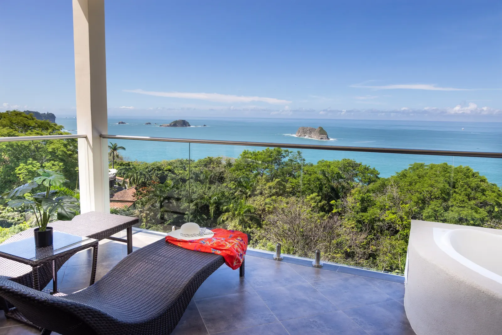 A hotel balcony with a wicker lounge chair, a sun hat, a corner jacuzzi tub, and a view of rocky islets in the ocean.