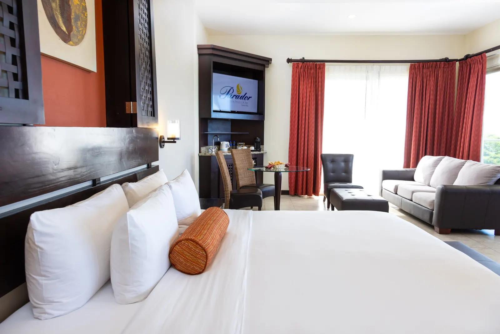 A bedroom with a dark carved wood headboard against a red wall, an orange accent bolster pillow, and dark leather and gray seating, with a framed painting partly visible on the adjacent wall.