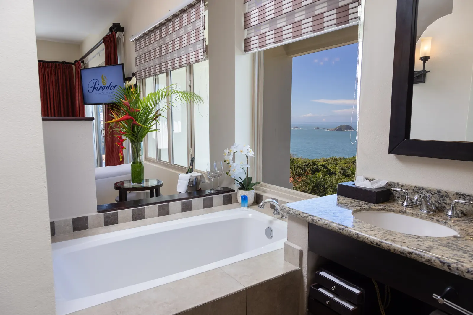 A soaking tub beneath windows overlooking the ocean, with flowers and a bottle of wine with two glasses on a nearby table.