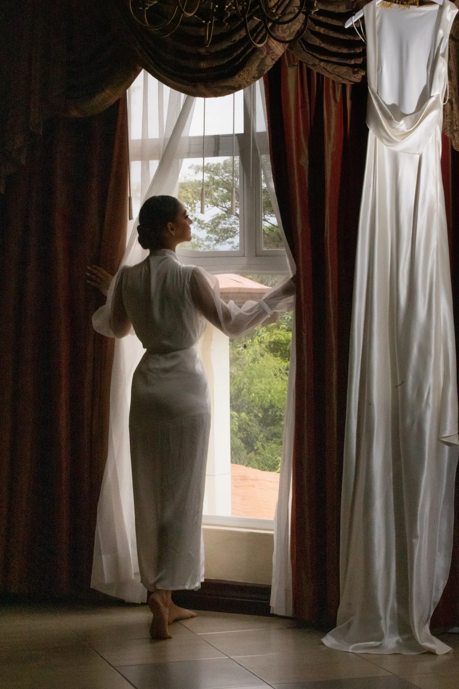 A bride in a white robe, seen from behind, opens the curtains of a suite window beside her hanging wedding gown.